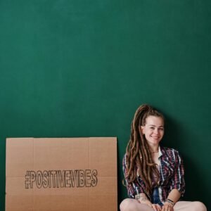 Smiling woman with dreadlocks sits next to a cardboard sign reading '#PositiveVibes'.